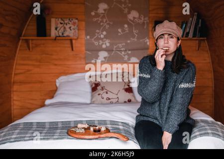 Blauäugige kaukasische junge Frau in Wollpullover und braunem Hut vor dem Essen einen appetitlichen herzhaften Snack mit kleinen Cerealien-Toasts und vor der Kamera Stockfoto