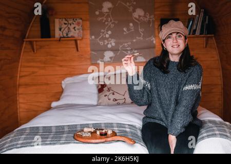 Blauäugiges kaukasisches Mädchen in Wollpullover und braunem Hut lächelt fröhlich der Kamera zu, die auf einem gemütlichen Bett in der Blockhütte sitzt, bevor sie einen kleinen Snack isst Stockfoto