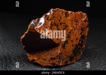 Echtes glänzendes Exemplar eines orangen Mahagoni Obsidian vulkanischen Steinmakros auf schwarzem Lederhintergrund isoliert Stockfoto