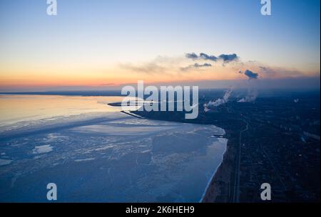 Luftaufnahme eines Industriewerks an der Küste eines gefrorenen Asowschen Meeres in Mariupol. Draufsicht auf die Fabrik, Rauch steigt aus den Schornsteinen. Stadt in der Nähe von Eis Stockfoto