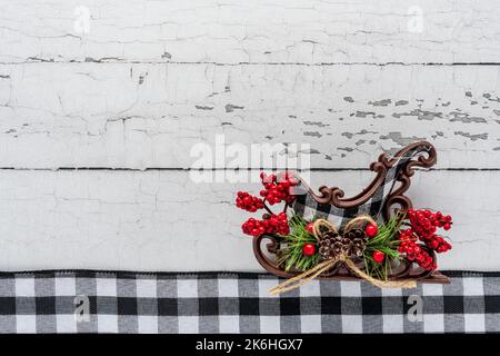 Winterurlaub-Thema mit schwarz-weißem Gingham-Band und einem Schlitten mit Kieferkegel und roten Beeren auf schälender Holzhintergrund Stockfoto