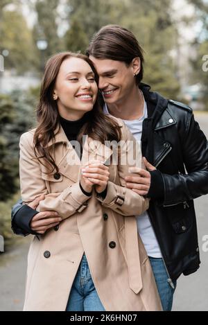 Fröhlicher junger Mann umarmt glückliche Freundin im Trenchcoat im Freien, Stockbild Stockfoto