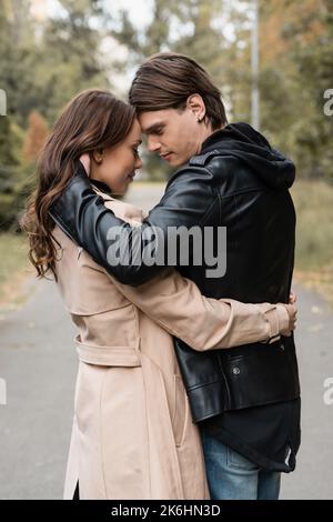 Seitenansicht des stilvollen jungen Mannes umarmt hübsche Freundin im Trenchcoat im Freien, Stockbild Stockfoto