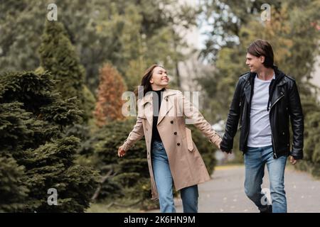Fröhliches und stilvolles Paar in herbstlichen Outfits, die beim Spazierengehen im Park die Hände halten, Stockbild Stockfoto