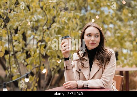 Verträumte Frau in beigem Trenchcoat halten Papiertasse mit Kaffee in herbstlichen Park gehen, Stock Bild Stockfoto