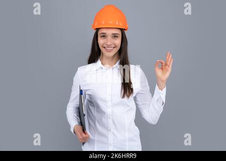 Lächelnde Frau in Schutzhelm mit Klemmbrett isoliert auf grauem Hintergrund. Junge Frau, Bauleiterin. Architektin Frau mit OK-Zeichen, weiblich Stockfoto