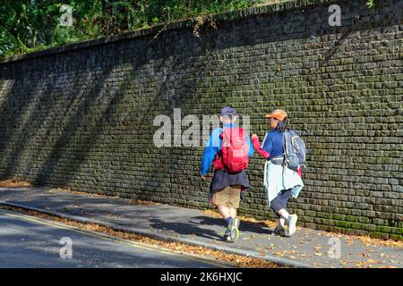 Ein orientalisches Paar mittleren Alters mit Wanderoutfit, das auf einem Bürgersteig mit einer großen Backsteinmauer im Hintergrund läuft, Shepperton Surrey England UK Stockfoto