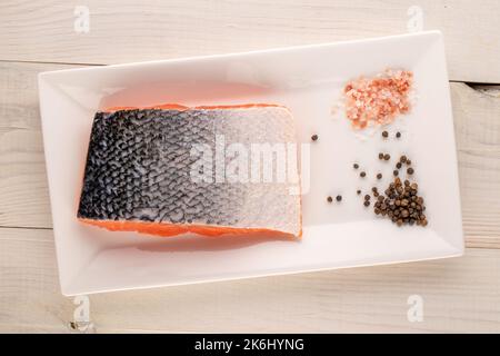 Ein Stück frisches Lachsfilet mit weißer Keramikplatte mit Pfeffer und Salz auf Holztisch, Makro, Draufsicht. Stockfoto