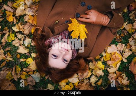 Porträt einer jungen rothaarigen Frau vor dem Hintergrund herbstlicher Blätter. Frau posiert, während sie auf dem Gras liegt und ein Flugblatt in der Hand hält Stockfoto