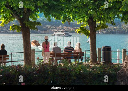 Italien Seen, Rückansicht einer Gruppe von Touristen mittleren Alters im Sommer sitzen auf einer Bank am See in Bellagio und beobachten Boote auf dem Comer See, Italien Stockfoto