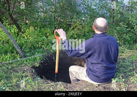 Der Mann gräbt eine tiefe Grube. Der Mensch ruht sich nach der Arbeit aus. Graben einer Grube durch Schaufel. Stockfoto