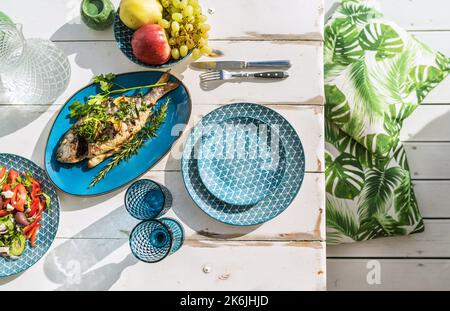 Im Ofen gebackene Goldbrasse, auch bekannt als Orata, wird auf einem weißen Holztisch mit Gemüsesalat und Obst serviert. Gesunde Zubereitung von Meeresfrüchten Stockfoto
