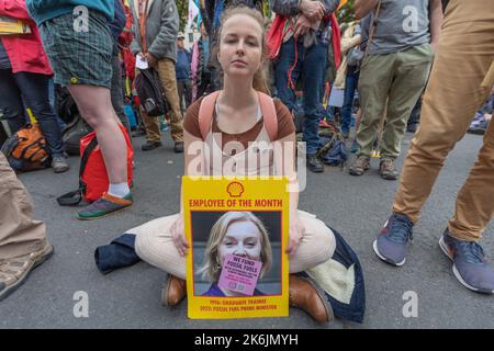 London, Großbritannien. Oktober 2022. Ein junger Demonstrant sitzt im Kreuz und hält ein satirisches Poster, das Shell's Corporate Branding parodiert. Das Poster zeigt ein Foto von Rt Hon Elizabeth Truss MP mit der Aufschrift „Shell Employee of the Month“ mit dem Slogan DOES FOSSIL FUEL PROUD MINISTER und einem pinkfarbenen Aufkleber mit der Aufschrift I AM A CLIMATE CRIMINAL. Die Szene fängt einen Klimaprotest mit Demonstranten und Plakaten im Hintergrund ein, der öffentliche Dissens gegen die Interessen fossiler Brennstoffe und politische Komplizenschaft hervorhebt. Penelope Barritt/Alamy Live News Stockfoto