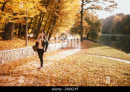 Glückliche junge Frau, die bei sonnigem Wetter im Herbst mit einem Smartphone einen Baum mit gelben Blättern fotografiert, während sie im Park spazieren geht Stockfoto