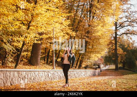 Glückliche junge Frau, die bei sonnigem Wetter im Herbst mit einem Smartphone einen Baum mit gelben Blättern fotografiert, während sie im Park spazieren geht Stockfoto