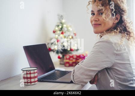 Seitenportrait der attraktiven jungen erwachsenen Frau, die lächelt und denkt, mit Laptop vor und weihnachtsschmuck im Hintergrund. Fröhliche Hündin Stockfoto