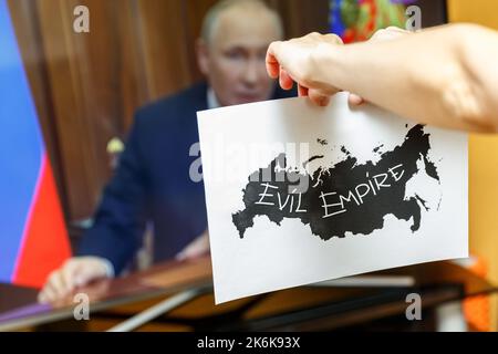 Hände zerreißen Papier mit schwarzen Konturen Russlands mit dem Ausdruck Böses Reich darauf, vor einem fernsehgerät. Russlands Zerfallskonzept Stockfoto