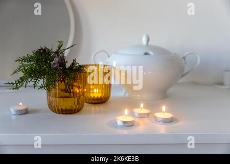 Gelbe Glasleuchter, ein Zweig von Arborvitae mit Zapfen, eine weiße Porzellanschüssel auf einer für Weihnachten dekorierten Kommode. Stockfoto