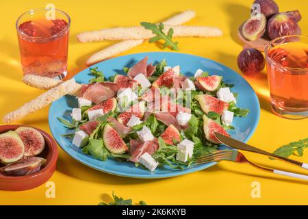 Herbstfeigen-Salat mit Rucola, Feta, rohem Schinken auf blauem Teller auf gelbem Hintergrund, serviert mit Rosenwein und Grissini. Flexitarian Diät, Paläo-Diät fallen Stockfoto