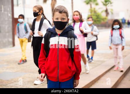 Schuljunge in medizinischer Maske in der Nähe der Schule, Kinder auf dem Hintergrund Stockfoto