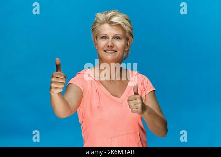 Lächelnde blonde Frau, die Daumen hoch gibt und ein pinkes Hemd trägt Stockfoto