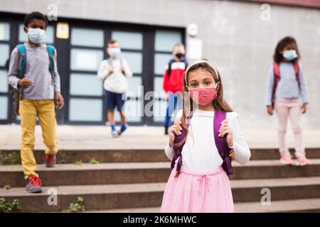 Schulmädchen in medizinischer Maske stehend auf der Straße, Kinder im Hintergrund Stockfoto