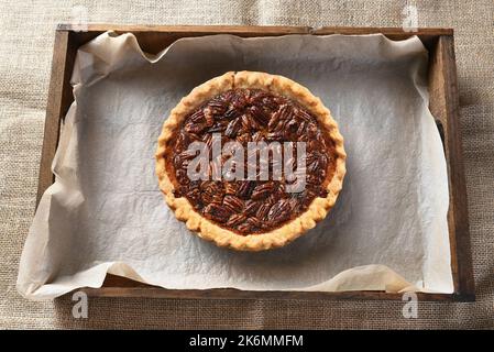 Ansicht eines Pecan-Kuchens in einer Holzkiste mit Pergamentpapier von oben. Der frisch gebackene Kuchen kühlt auf einem Tisch mit einem Lappen-Tischtuch ab. Stockfoto