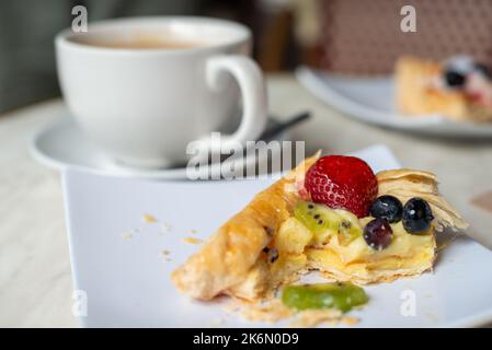 Eine weiße Porzellantasse und Untertasse mit einem kleinen Teelöffel auf der Untertasse. Der Becher ist mit heißem Kaffee und Creme gefüllt. Der Teller hat ein Gebäck mit Beeren. Stockfoto