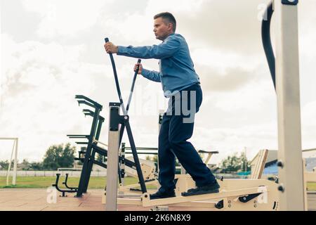 Ein Mann in einem Geschäftsanzug wärmt sich auf Simulatoren im Stadion auf. Sportübungen nach der Büroarbeit. Der Kampf gegen Übergewicht. Geschäftsmann Stockfoto