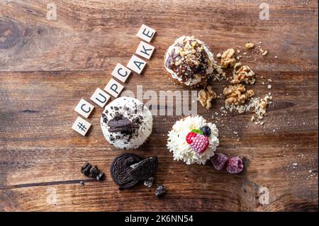 Drei leckere Cupcakes auf einem Holztisch. Weiße Creme, Schokolade und Himbeere, mit Nuss überzogen. Das Wort Cupcake aus Holzblöcken. Stockfoto