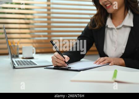 Lächelnde junge Geschäftsfrau, die in ihrem persönlichen Büro sitzt und Dokumente unterschreibt. Zugeschnittenes Bild Stockfoto