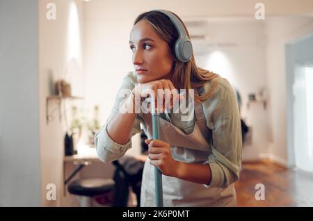 Frau, müde und putzte mit Kopfhörern für Musik, während sie im Zimmer zu Hause arbeitete. Sauberer, denkender und nachdenkender beim Hören von Radio, Podcast Stockfoto