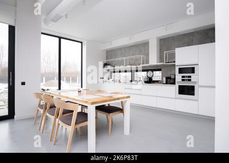 Elegante weiße Küche mit einem großen Panoramafenster ohne Menschen. Stilvoller Holztisch in der Küche. Frontansicht Banner eines Küchentisches Stockfoto
