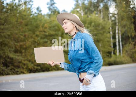 Junge Frau schaut hoffentlich vorbei an Autos mit leerem Pappplakat am Straßenrand im Wald. Lady im Hut- und Denim-Outfit entfliehen Sie der Stadt Stockfoto