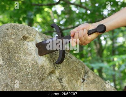 Hand des Ritters, der versucht, das Excalibur-Schwert zu extrahieren, das in den Felsen mitten im verzauberten Wald stecken geblieben ist Stockfoto