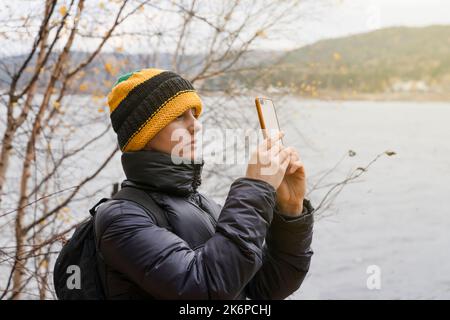 Eine Frau in einer warmen Jacke fotografiert die Natur im Spätherbst. In einem gelben Hut gekleidet, macht Fotos von der Natur und den Wäldern am Fluss oder See im Freien Stockfoto