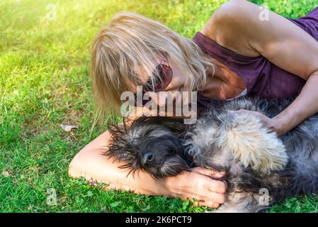 Nahaufnahme einer blonden Frau mittleren Alters, die auf dem Gras liegt, mit ihrem katalanischen Schäferhund, der spielt und kuschelt. Liebe für Haustiere. Adoption von Tieren Stockfoto