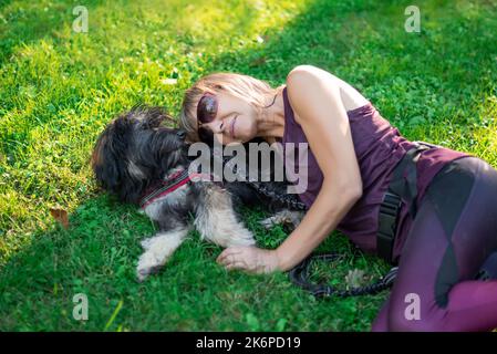 Eine blonde Frau mittleren Alters, die auf dem Gras liegt und sich auf den Rücken ihres katalanischen Schäferhundes lehnt, der sich ausruht und die Gesellschaft ihres Haustieres genießt. Liebe für Stockfoto