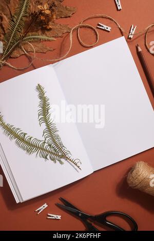 Getrocknetes gepresstes Blatt auf Papier aus Notizbuch mit Bleistift, Schere und Garn auf braunem Hintergrund. Hobby, handgemachte, florale Kunst und Boho Style Konzept Stockfoto