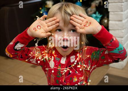 Kind in weihnachten roten Schlafanzug und Hirsch Make-up auf Gesicht gemalt macht lustige Gesichter. Die Zahlen 2023 sind auf Palmen geschrieben. Weihnachten, Neujahr, fröhlicher Winter Stockfoto