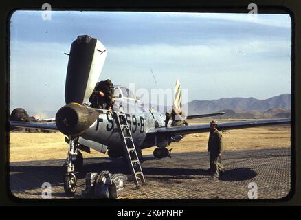 Dreiviertel linke Vorderansicht eines Republic F-84 Thunderjet 'This'll Kill Ya' (s/n 46-559) mit den Markierungen des 8. Fighter Bomber Squadron (FBS), 49. Fighter Bomber Group (FBG), irgendwo in Korea. Das Nasenfeld ist offen und ein Bodenbemannter führt Wartungsarbeiten durch. Ein anderer Mann arbeitet am Flügel, und ein dritter wird gezeigt, wie er in Richtung des Flugzeugs geht. Eine Leiter wird gegen die Seite des Flugzeugs aufgesetzt, die zum Cockpit führt. Einige Geräte werden am Vorderrad platziert. Im Hintergrund ist mehr Ausrüstung zu sehen. Stockfoto