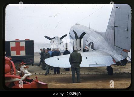 Dreiviertel linke Seitenansicht eines Douglas C-47B Skytrain (s/n 43-48930; c/n 14747-26191)-Flug mit dem Flugzeug, irgendwo in Korea. Ein Krankenwagen wird links vom Flugzeug geparkt, und eine Gruppe von Männern steht neben dem Flugzeug in der Nähe der Flugzeugtür, die geöffnet ist. In der unteren linken Ecke des Bildes wird die Stützausrüstung angezeigt. Stockfoto