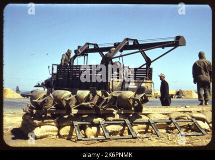 Eine halbe linke Rückansicht eines LKW mit Winde und Riemenscheibe zum Be- und Entladen von Bomben, irgendwo in Korea. Ein Mann steht zur Vorderseite des Lacks, während ein anderer in der Mitte steht, um Bomben zu laden und zu entladen. Zwei weitere Männer stehen hinter dem LKW. Fünf Bomben sind Bilder im Vordergrund, die auf einem niedrigen Stapel von Sandsäcken platziert sind. Im Hintergrund ist ein Lockheed F-80 Shooting Star zu sehen, sowie ein LKW und zwei Umbauten. Stockfoto