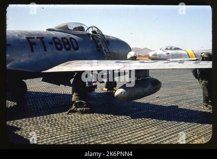 Eine halbe rechte Rückansicht eines Lockheed F-80C Shooting Star (s/n 49-680) mit den Markierungen des 36. Fighter Bomber Squadron (FBS) irgendwo in Korea. Der rechte Flügel ist mit einem Napalmkanister ausgestattet und unter dem Flügel sitzt ein Bodencrewman, der eine Rakete befestigt. Ein weiterer Mann steht vor dem Flügel, rechts vom Bild. Ein nordamerikanischer Säbel der F-86 des 4. Fighter Interceptor Wings (FIW) steht direkt vor der F-80. Stockfoto