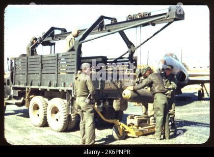 Dreiviertelansicht links von hinten eines LKW, der mit einer Winde und einem Riemenscheibensystem zum Be- und Entladen von Bomben irgendwo in Korea ausgestattet ist. Zwei Männer stehen im LKW, drei weitere stehen dahinter, laden oder entladen eine Bombe. Vor ihnen sitzt ein Wagen für den Transport der Bombe auf dem Boden. Ein Lockheed F-80 Shooting Star ist senkrecht zum LKW geparkt. Im Hintergrund erscheinen Gebäude und Sandsäcke. Stockfoto