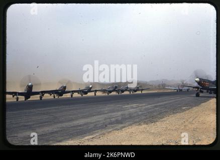 Dreiviertelansicht von rechts hinten eines nordamerikanischen F-51D Mustang, der irgendwo in Korea rollt. Neben der Start- und Landebahn auf der linken Seite des Bildes ist eine Fluglinie von F-51D Mustangs geparkt. Ein weiterer F-51D Mustang ist auf der rechten Seite der Start- und Landebahn abgestellt. Drei der Flugzeuge tragen die Markierungen des 12. Fighter Bomber Squadron (FBS). Jedes Flugzeug ist mit zwei Bomben bewaffnet. Stockfoto