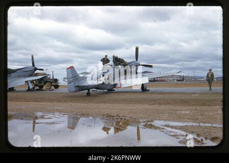 Eine halbrechte Rückansicht eines nordamerikanischen RF-51D Mustang (s/n 44-84775; c/n 44631), der irgendwo in Korea geparkt ist. Auf dem linken Flügel stehen ein Schiffsführer und der Pilot. Ein weiterer Mann steht auf dem rechten Flügel. Ein zweiter RF-51D Mustang und ein Jeep sind auf der linken Seite des Flugzeugs geparkt. Zwei Convair PB4Y-2 Privateer von VP-871 und ein Douglas R-4D1 Skytrain erscheinen im Hintergrund. Stockfoto