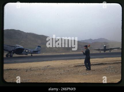 Ein Viertel von links Vorderansicht eines nordamerikanischen F-51D Mustang (s/n 44-73860; c/n40400), der irgendwo in Korea rollt. Ein weiterer F-51D Mustang ist neben der Start- und Landebahn abgestellt. Beide sind mit zwei Bomben bewaffnet. Im Vordergrund erscheint ein Besatzungsmann mit einer Flagge. Stockfoto