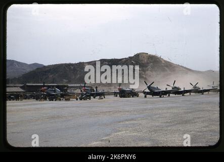 Eine halbe linke Vorderansicht eines nordamerikanischen F-51D Mustang, der irgendwo in Korea auf einer Fluglinie mit vier anderen ähnlichen Flugzeugen geparkt ist. Jede ist mit zwei Bomben bewaffnet. Rechts von der Fluglinie befinden sich drei nordamerikanische Mustang Mk. IVS mit den Markierungen der südafrikanischen Luftwaffe. Bodencrewmen, Unterstützungsfahrzeuge und Ausrüstung umgeben sie, die Betankung und Wartungsarbeiten durchführen. Im Hintergrund sind mehrere Gebäude zu sehen. Stockfoto