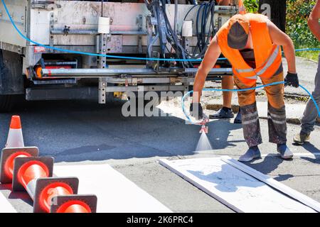 Ein Straßenarbeiter, der einen Farbsprüher verwendet, trägt weiße Straßenmarkierungen auf einen gestreiften Fußgängerüberweg mit einer Holzschablone auf. Speicherplatz kopieren. Stockfoto